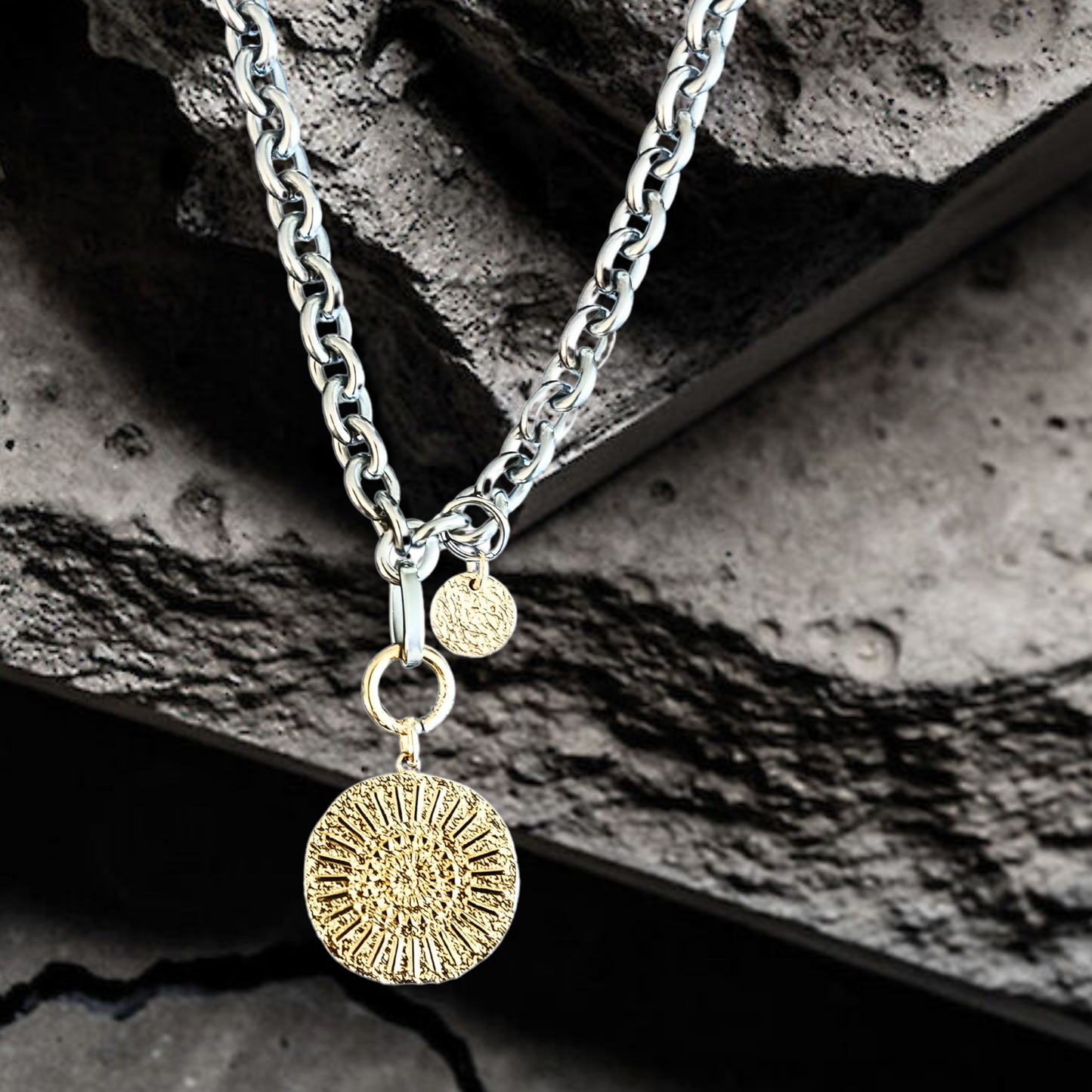 Moderne silberfarbene Halskette aus Edelstahl mit einem goldplattierten Münzanhänger. Der Anhänger ist 23mm groß und hat ein abstraktes Hammerschlagmuster mit Strahlen. Handgefertigter Edelstahlschmuck der Schmuckmarke Hug the Lioness aus Österreich. Die Kette ist 6mm breit. Neben dem Karabiner ist ein 10mm vergoldetes, gehämmertes Plättchen aus Sterlingsilber angebracht.