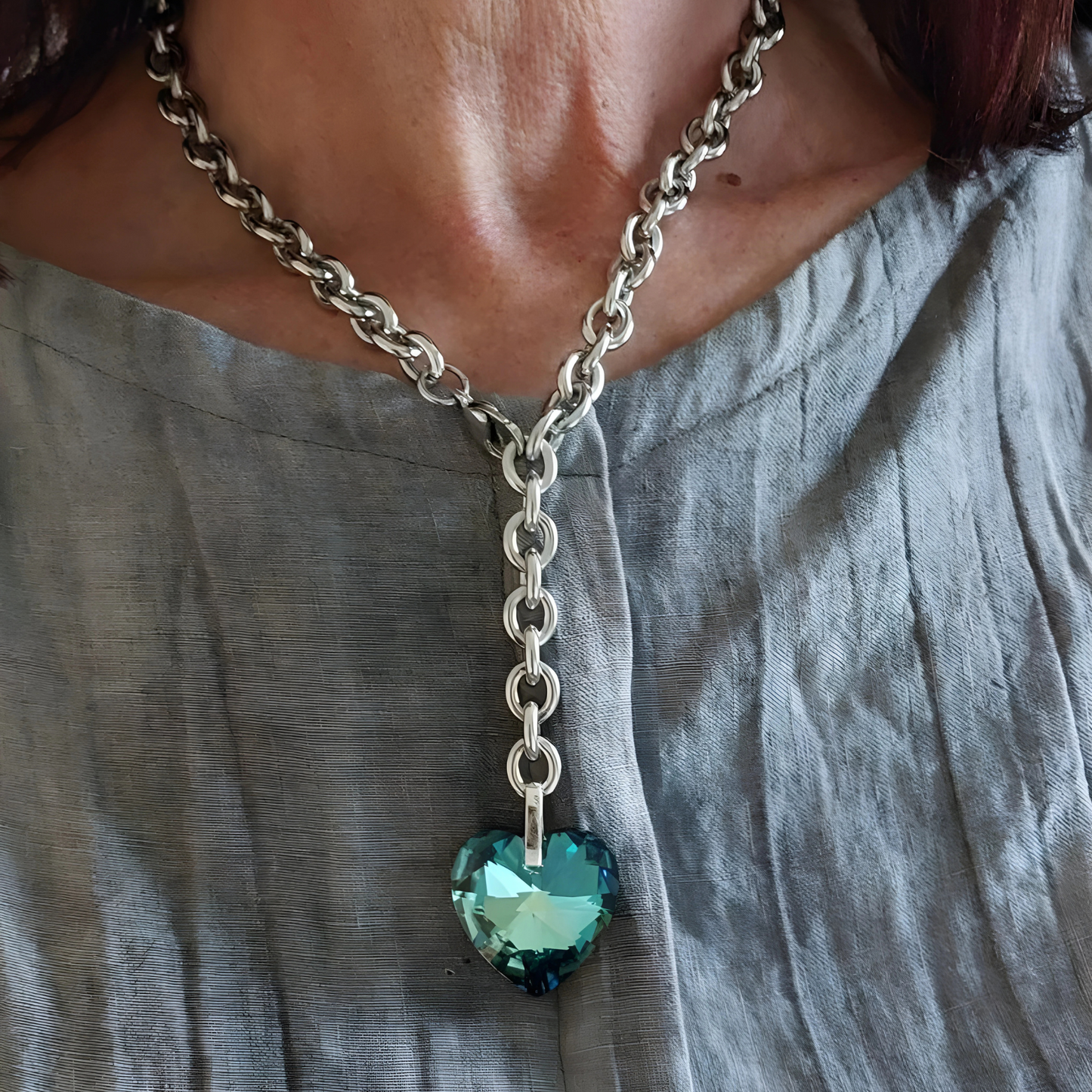 Titanic Halskette aus silberfarbenem Edelstahl mit einem großen blauen Kristallherz. Die Kette hat 9mm breite Kettengliedern. Der 28mm große Kristallanhänger wurde in Österreich gefertigt. Die Kette ist mit einem Karabiner vorne zu schließen und kann normal oder als Y-Kette getragen werden. Edelstahlschmuck von Hug the Lioness aus Österreich. Handgefertigt in Österreich. Die Halskette wird getragen von einer Frau im grauen Top.