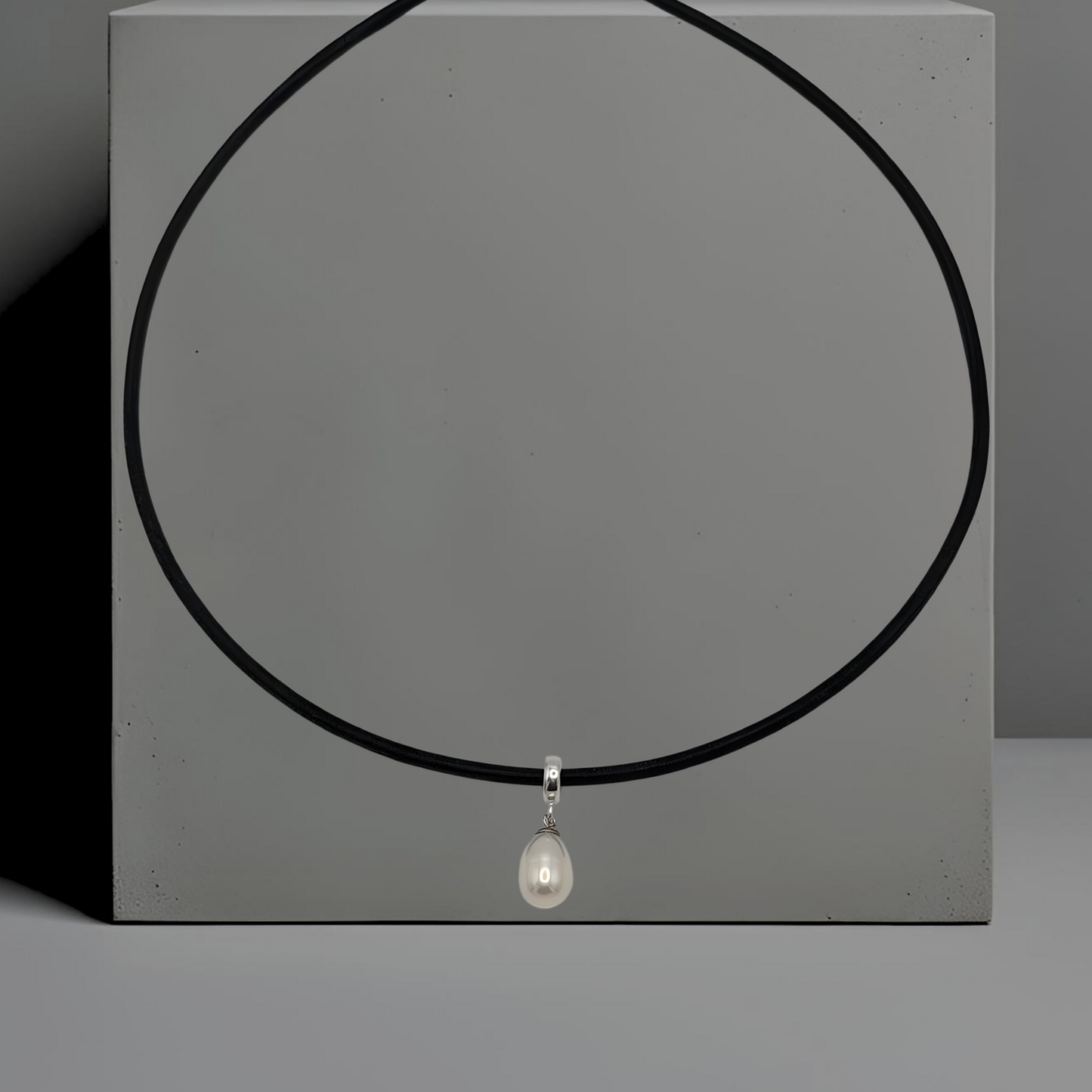 Schwarze 45cm Lederkette, rund, mit einer weißen Süsswasserperle als Anhänger. Die längliche Perle ist 9*11 mm gross und mit einem Anhängerring aus Silber an der Lederkette angebracht. Die Kette hat einen Karabinerverschluss. Der Hintergrund der Kette ist grau.