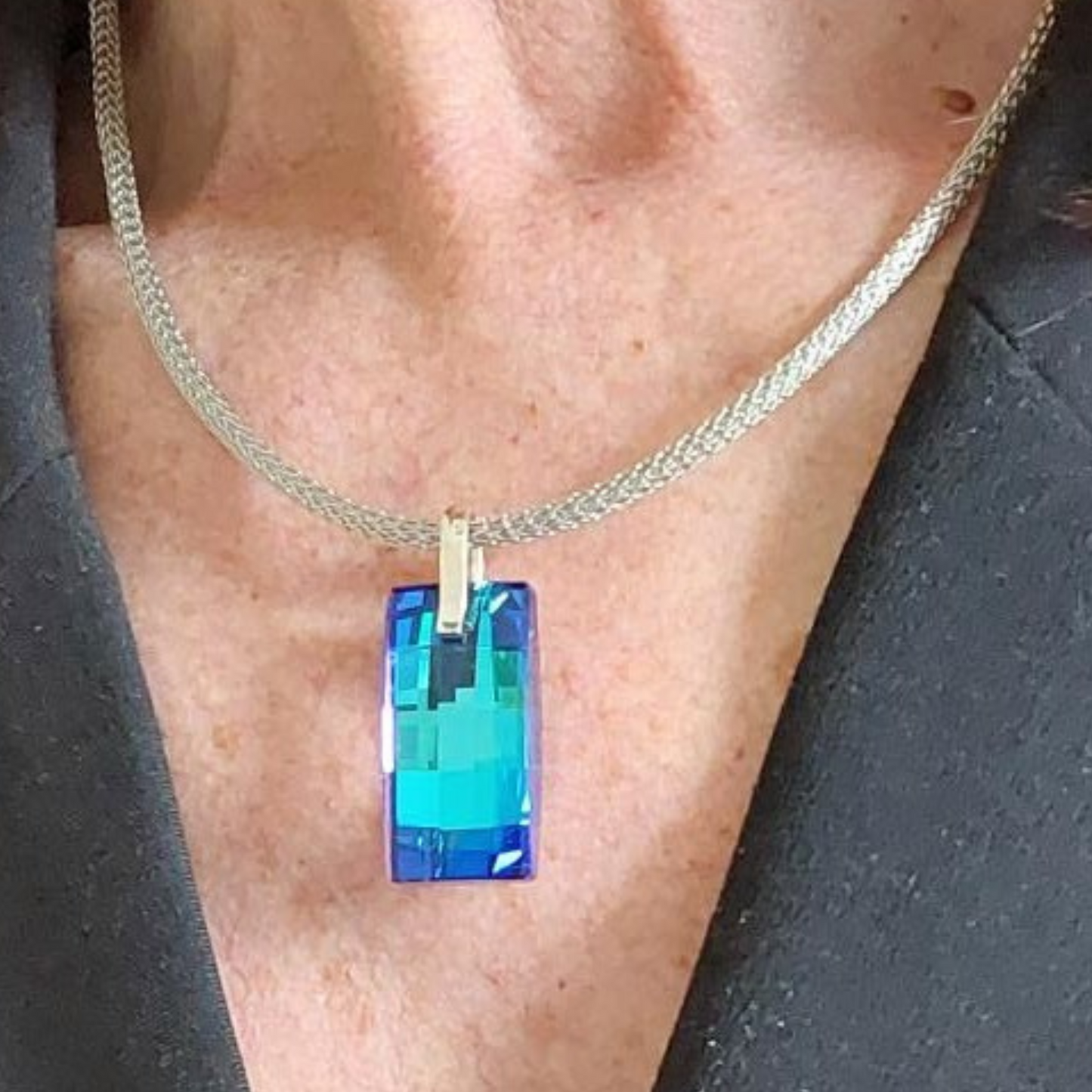 Silberfarbene Mesh Halskette aus Edelstahl mit einem großen rechteckigen blauen Kristallanhänger. Die moderne Meshkette ist 3mm breit und 45cm lang und stabil wie ein Halsreif. Der Kristall Urban ist 8mmx14mm in der Farbe Bermuda Blue. Der Kristall wurde in Österreich hergestellt. Die Kette hat einen Bajonettverschluss. Die Halskette wird von einer Frau im schwarzen Blazer getragen.