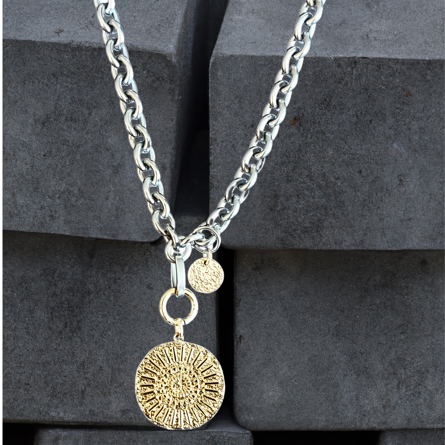 Goldplattierter Münzanhänger an einer breiten Gliederkette aus silberfarbenem Edelstahl . Die Münze ist 23mm und hat ein abstraktes Hammerschlagmuster mit Strahlen. Handgefertigter Edelstahlschmuck der Schmuckmarke Hug the Lioness aus Österreich. Die Kette ist 6mm breit. Neben dem Karabiner ist ein 10mm vergoldetes, gehämmertes Plättchen aus Sterlingsilber angebracht.