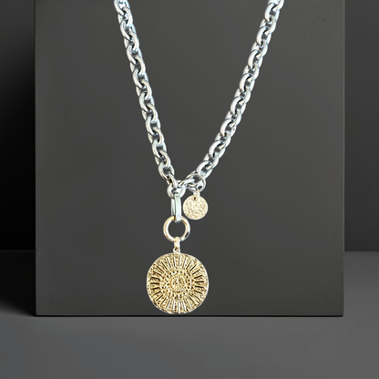 Moderne silberfarbene Halskette aus Edelstahl mit einem goldplattierten Münzanhänger. Der Anhänger ist 23mm groß und hat ein abstraktes Hammerschlagmuster mit Strahlen.  Handgefertigter Edelstahlschmuck der Schmuckmarke Hug the Lioness aus Österreich. Die Kette ist 6mm breit. Neben dem Karabiner ist ein 10mm vergoldetes, gehämmertes Plättchen aus Sterlingsilber angebracht.