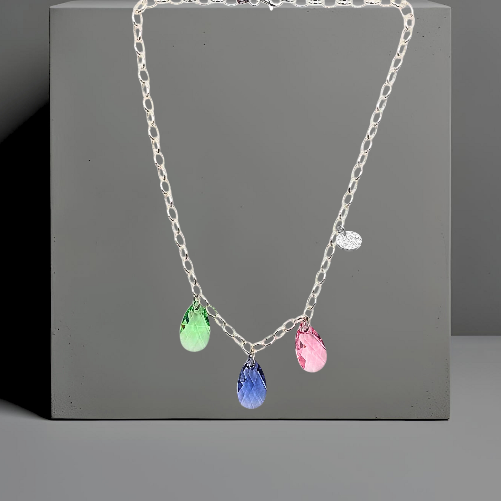 Collier aus 925 Sterlingsilber,  Gliederkette 5mm vorne mit 3 Kristalltropfen in rose, violett und grün. Die Kristalle sind aus Österreich. Die Kette ist 42cm lang und hat einen Karabinerverschluss. An der Seite ist in kleines gehämmertes Plättchen aus 925 Sterlingsilber angebracht. Die Kette ist aus der Kollektion JOY der Schmuckmarke HUG THE LIONESS.