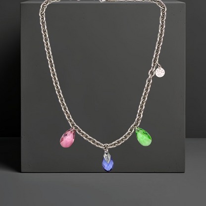 Collier aus Edelstahl, silberfarbene Gliederkette vorne mit 3 Kristalltropfen in rose, violett und grün. Die Kristalle sind aus Österreich. Die Kette ist 43cm lang und hat einen Karabinerverschluss. An der Seite ist in kleines gehämmertes Plättchen aus 925 Sterlingsilber angebracht. Die Kette ist aus der Kollektion JOY der Schmuckmarke HUG THE LIONESS.