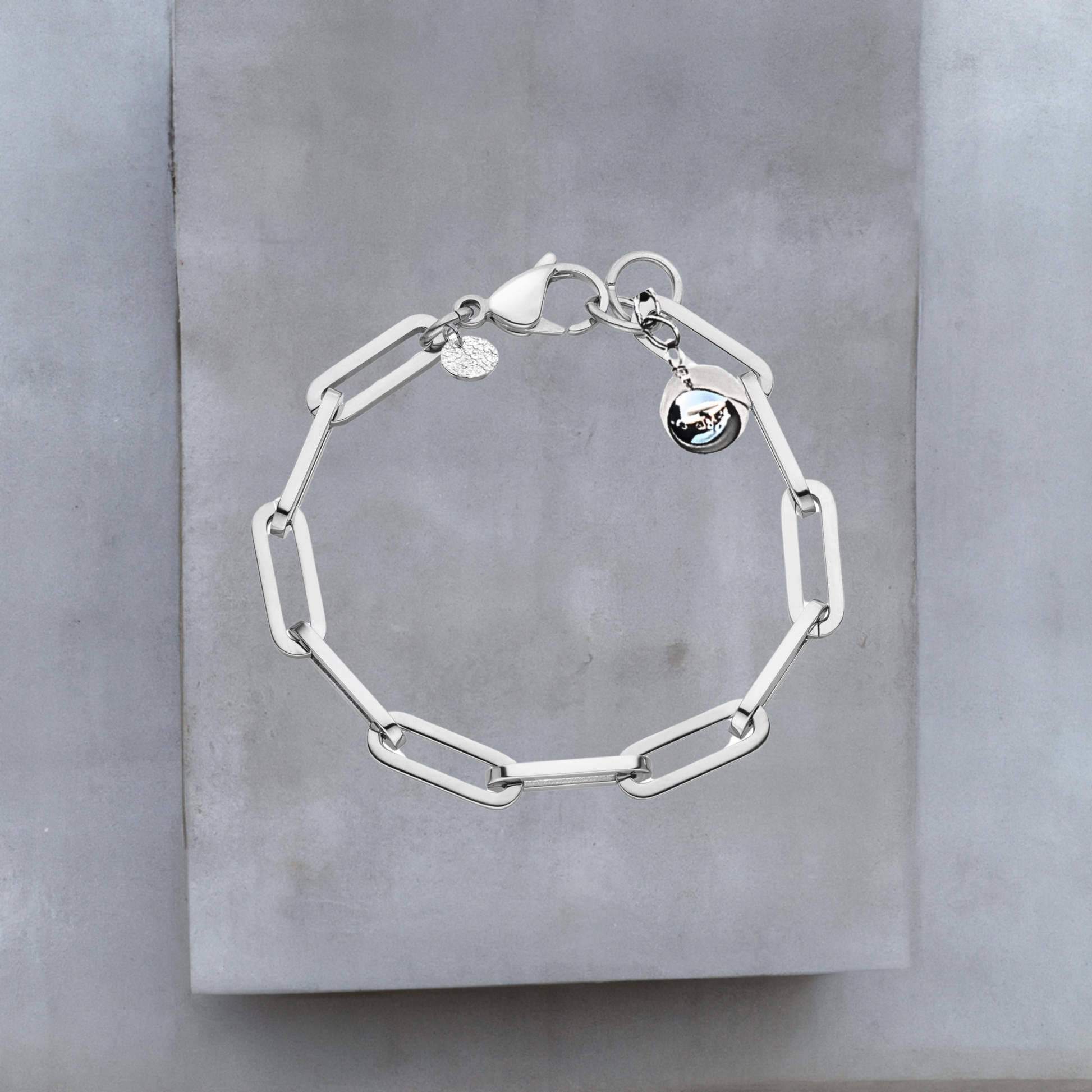Exklusive Armkette von Hug the Lioness Schmuck. Silberfarbene Armkette aus glattem Edelstahl mit weiten Kettengliedern. An einem Ende der Armkette ist eine glänzende silberne Kugel aus Sterlingsilber angebracht. Neben dem großen Karabinerverschluss ist ein kleines gehämmertes Silberplättchen angebracht.