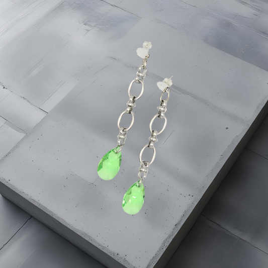 Topmodische 8cm lange Ohrhänger aus rostfreiem Edelstahl bestehend aus einer Kette mit 10mm breiten länglichen Kettengliedern und einem 22mm großen Kristalltropfen am Ende der Kette in peridot grün. Am oberen Ende ist ein Silberplättchen mit einen Steckverschluss angebracht. Die Ohrhänger sind ausdrucksstark und modern. Der Ohrschmuck ist von der österreichischen Schmuckmarke Hug the Lioness und handgemacht. Die Kristalle sind in Österreich hergestellt.