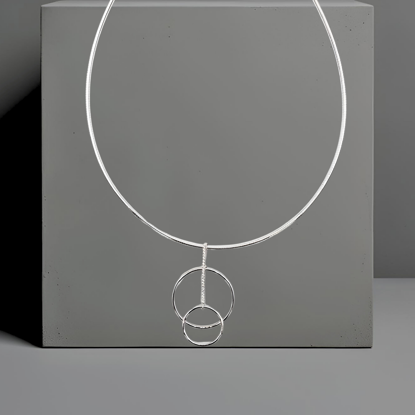 Feiner Omegareif Lara mit Anhänger. Halskette Material: 925 Sterlingsilber
Länge: 42 cm, Stärke: 1,9mm
Verschluss: Karabiner.
Der zierliche Anhänger besteht aus 2 versetzten Kreisen. Die Kreise sind durch einen schmalen Steg verbunden. Der Steg ist mit 20 kleinen weißen Zirkonia besetzt. Der Anhänger ist 29,8mm breit und 50,7mm hoch. 