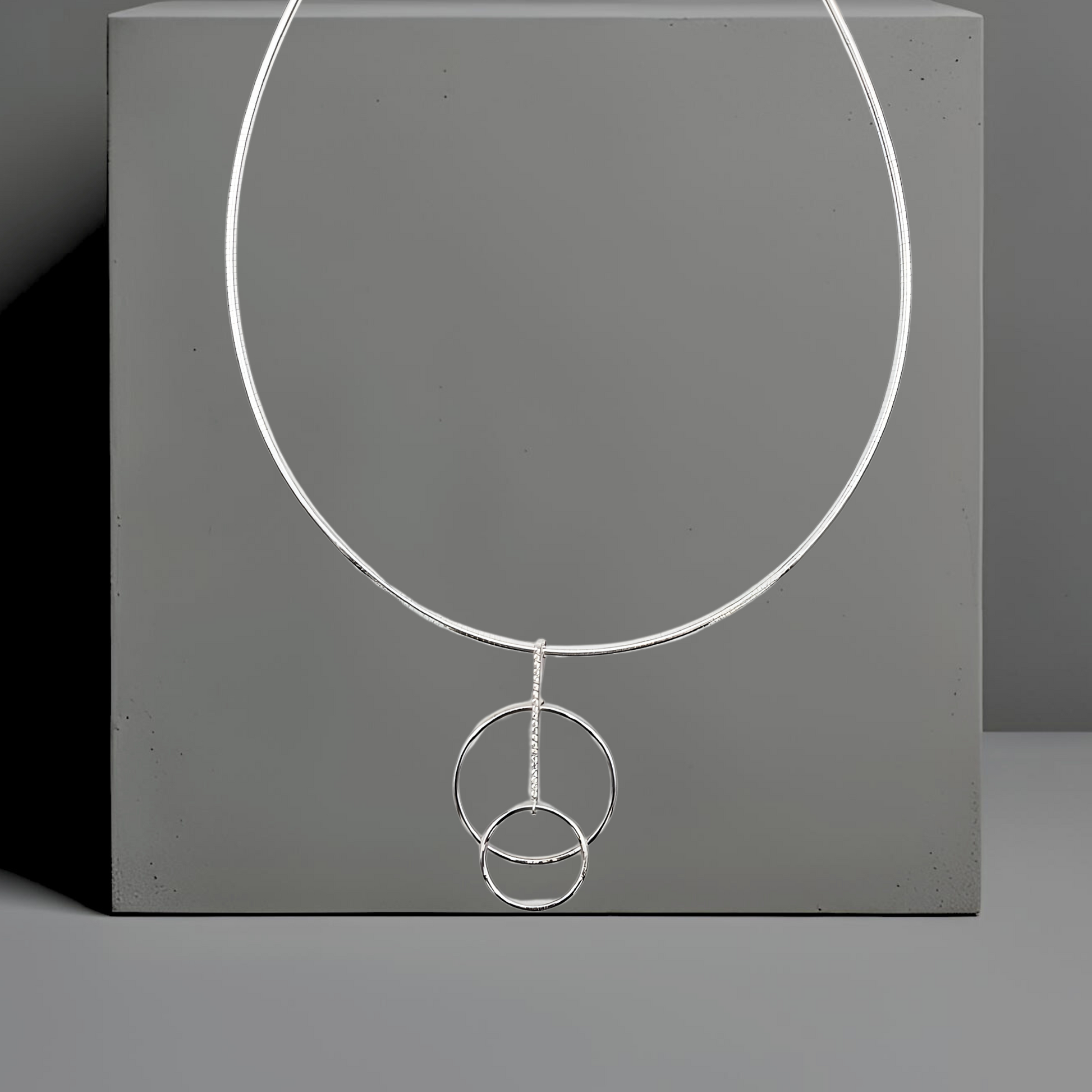 Feiner Omegareif Lara mit Anhänger. Halskette Material: 925 Sterlingsilber
Länge: 42 cm, Stärke: 1,9mm
Verschluss: Karabiner.
Der zierliche Anhänger besteht aus 2 versetzten Kreisen. Die Kreise sind durch einen schmalen Steg verbunden. Der Steg ist mit 20 kleinen weißen Zirkonia besetzt. Der Anhänger ist 29,8mm breit und 50,7mm hoch. 