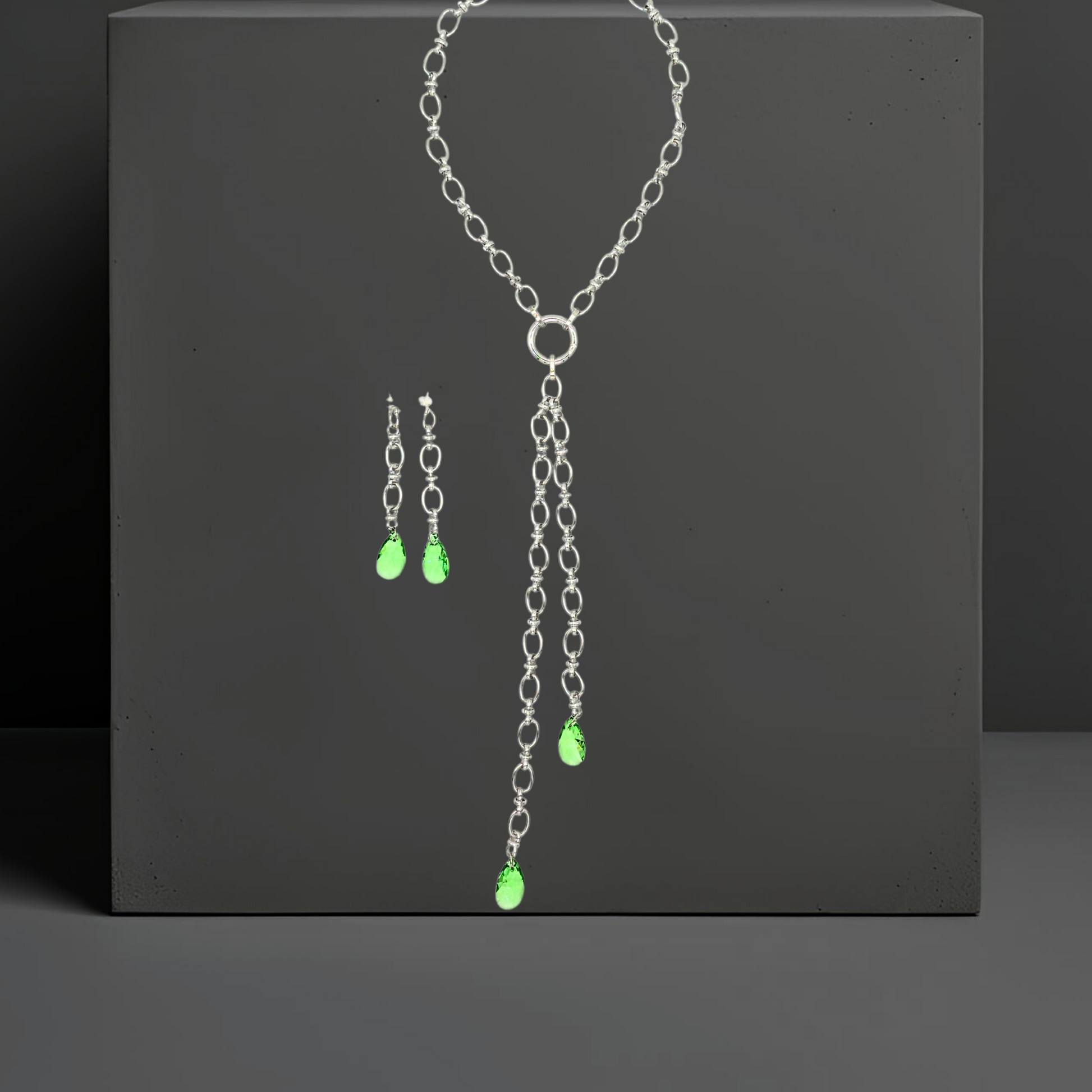 Handgefertigte breite Gliederkette aus rostfreiem Edelstahl mit einem Ringverschluss vorne. Die passenden Ohrhänger sind ebenfalls abgebildet. Im Ringverschluss der Kette ist ein langer Anhänger angebracht, der aus 2 Ketten (17cm und 25cm) und jeweils einem peridot grünem Kristalltropfen am Ende der Ketten besteht. Der lange Anhänger kann auch abgenommen werden und die Kette mit dem Ringverschluss als kurzes Collier getragen werden. Die Halskette ist von der Schmuckmarke Hug the Lioness aus Österreich. 